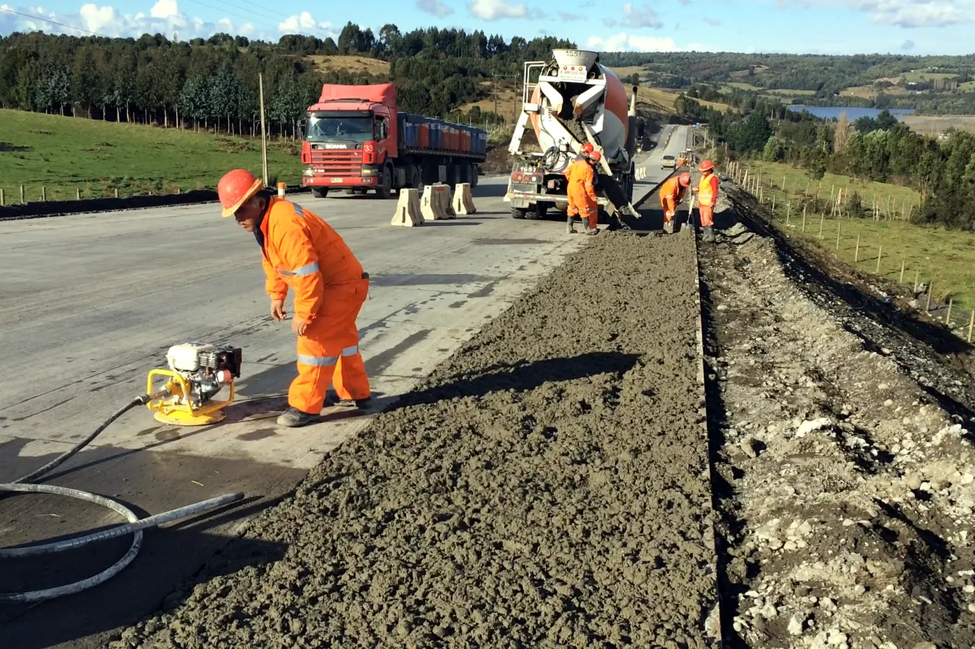 Un operario compactando la berma junto a la calzada de hormigón recién extendida, con los camiones al fondo