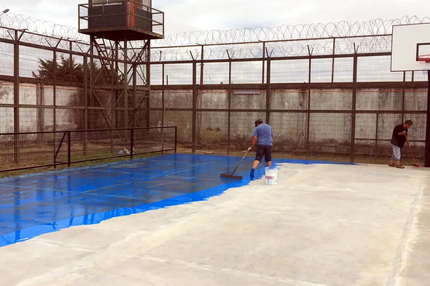 Aplicación del recubrimiento azul sobre la losa de una cancha deportiva, con el cierre perimetral y el aro montados