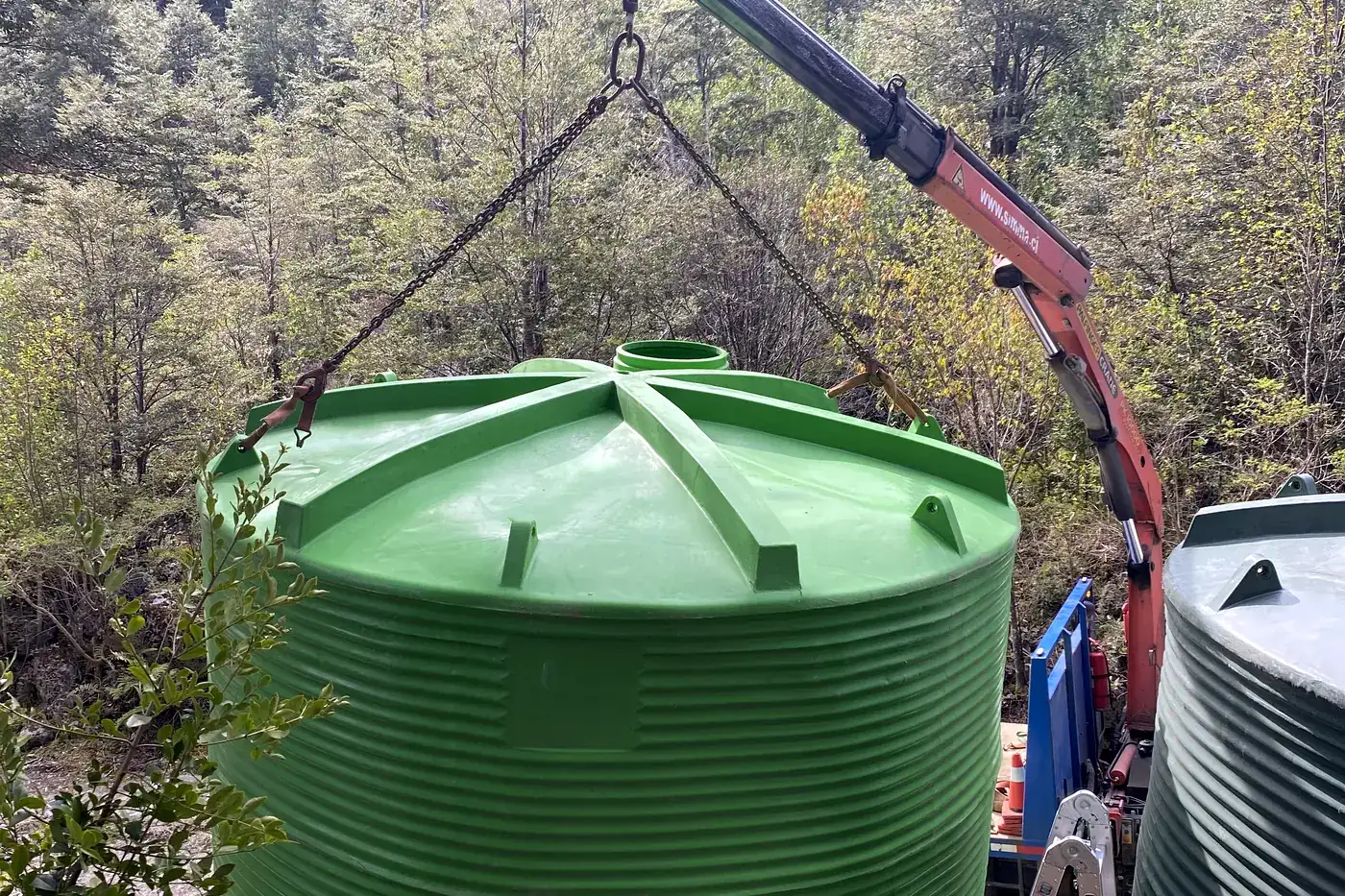 Un estanque de agua suspendido del brazo de un camión grúa sobre su radier, con el bosque nativo alrededor