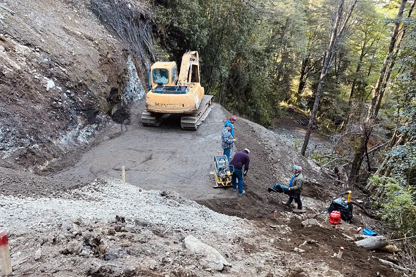 Excavadora sobre la plataforma de un camino de montaña, con la cuadrilla trabajando al costado