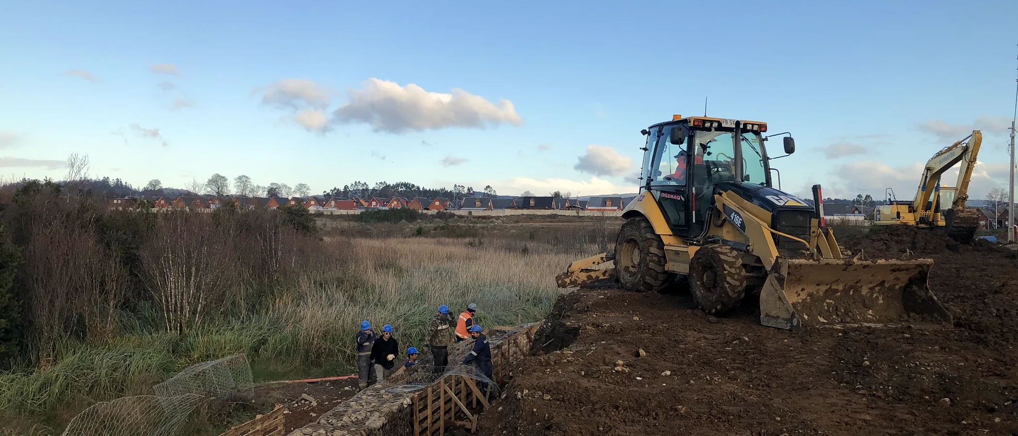Faena de contención de gaviones junto a un humedal: la cuadrilla armando la canastilla de malla al pie de la excavación, una retroexcavadora sobre el terraplén y una excavadora al fondo, con el barrio y la línea de árboles en el horizonte